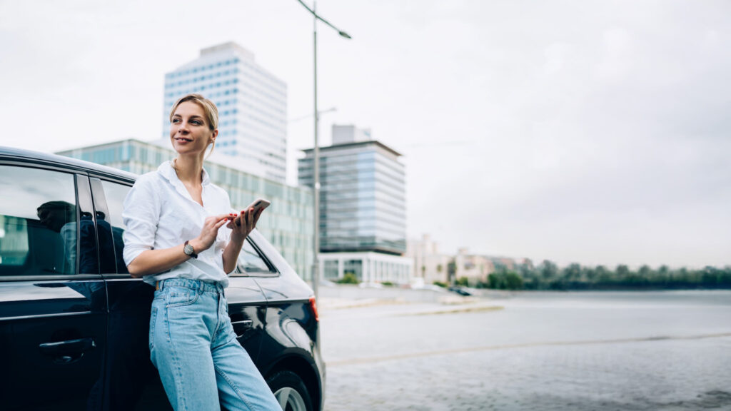 Person lehnt an Auto am Wasser und nutzt Smartphone